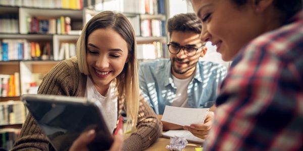 Close up of three smiling lovely playful young students sitting in the high school library and looking in a tablet while sitting at the desk together.