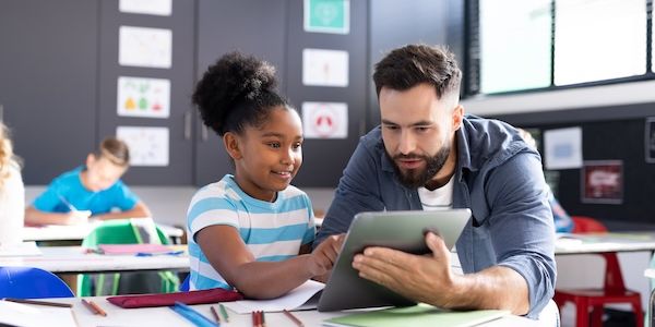 Diverse male teacher and happy girl using tablet at her desk in elementary classroom 