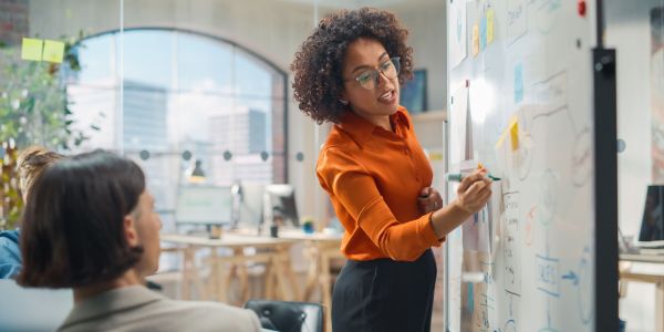 Diverse Modern Office: Black Businesswoman Leads Business Meeting with Managers, Talks, Explains Statistics, Uses a Whiteboard with Graphs, Big Data. Digital Entrepreneurs Work on a Project 